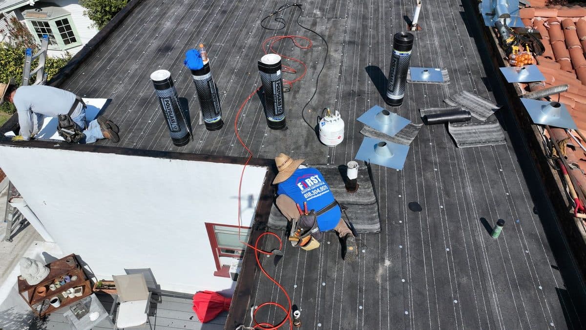 Aerial view of crew members working on a dark flat roof with pipe vent flashings and propane tanks