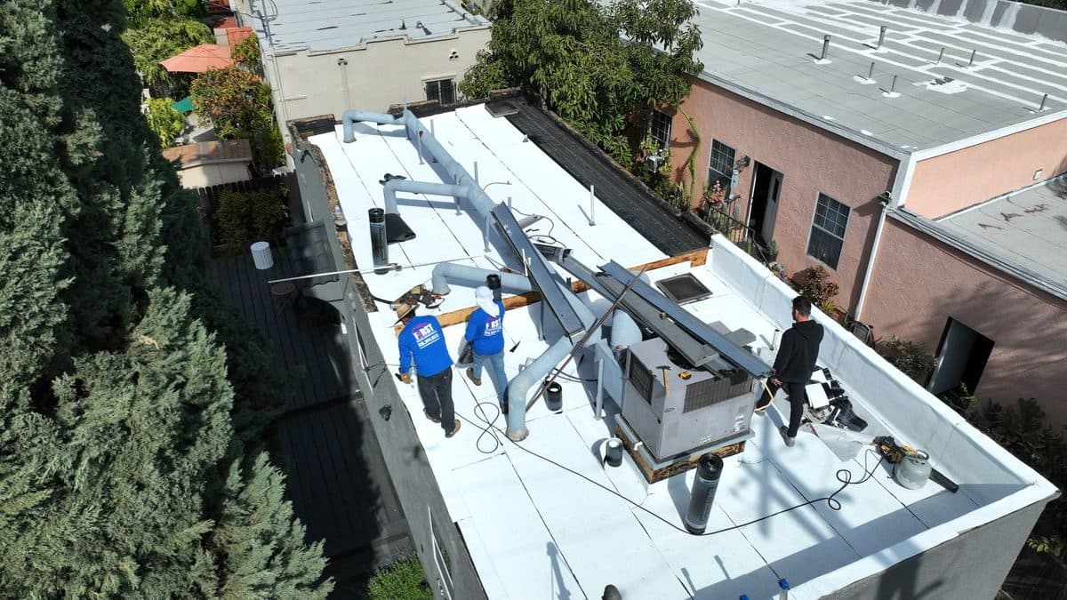 Aerial view of crew members and a person in black beside rooftop equipment on a white flat roof