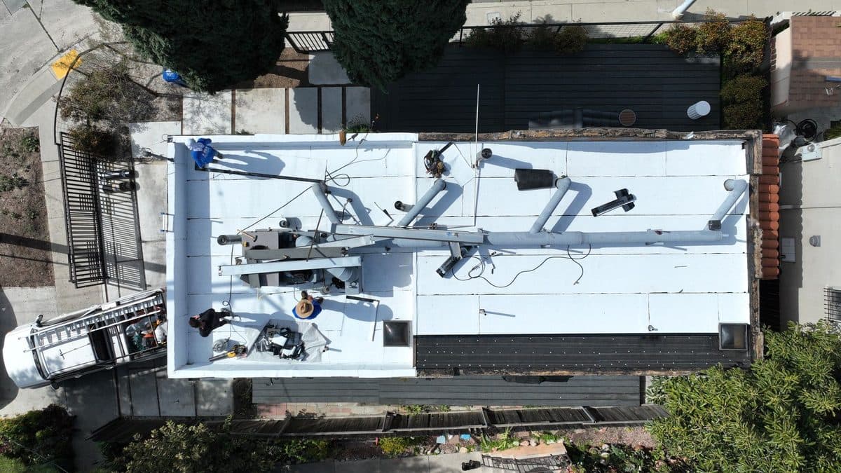 Top-down aerial view of a white flat roof with rooftop piping, a ladder and a work truck