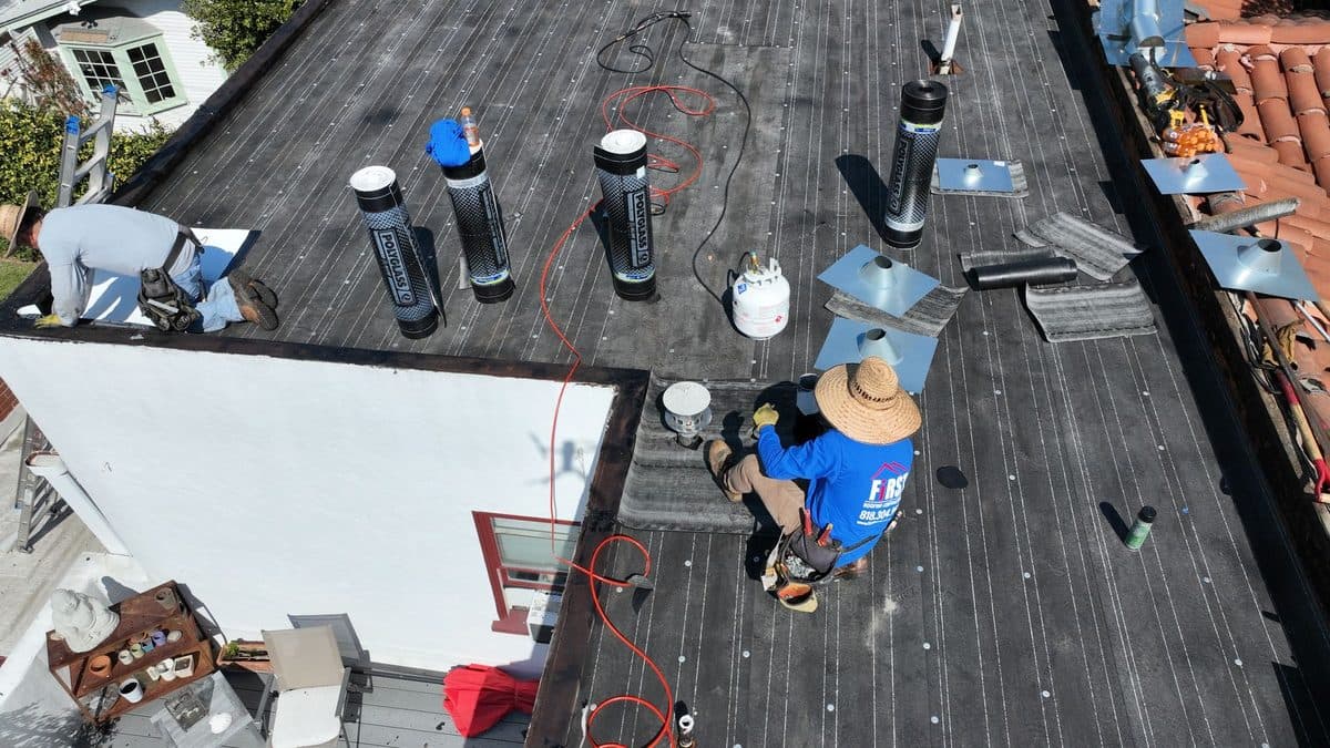 Close aerial view of a crew member working on a dark flat roof with pipe vents and propane tanks
