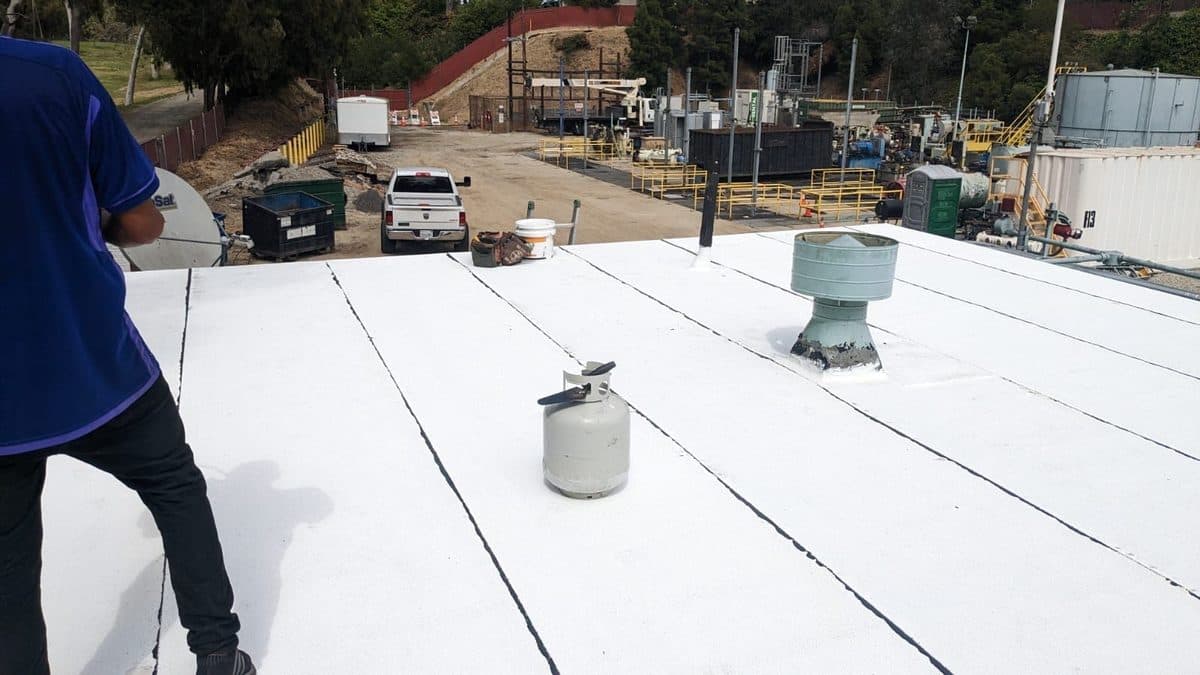 Crew member standing on a white flat roof beside a propane tank and a roof vent