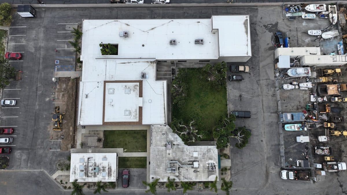 Top-down aerial view of a white-roofed commercial building complex beside a yard