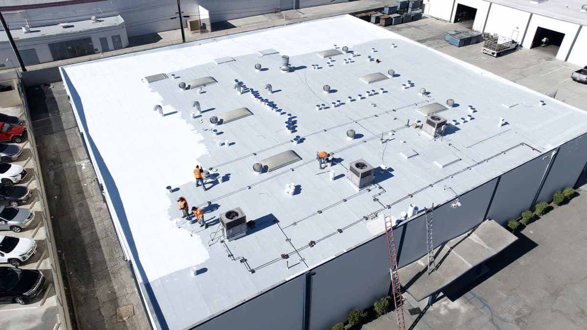Aerial view of crew members coating a large commercial flat roof, with part of the surface still gray