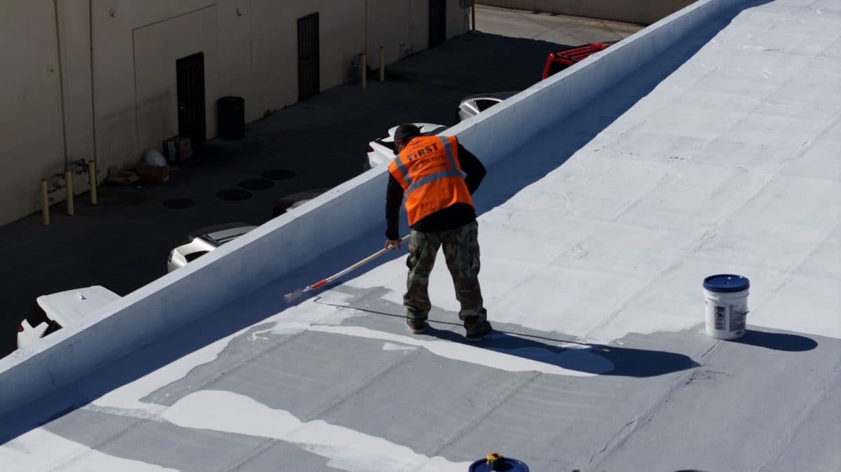 Crew member in an orange vest working on a white flat roof beside a bucket