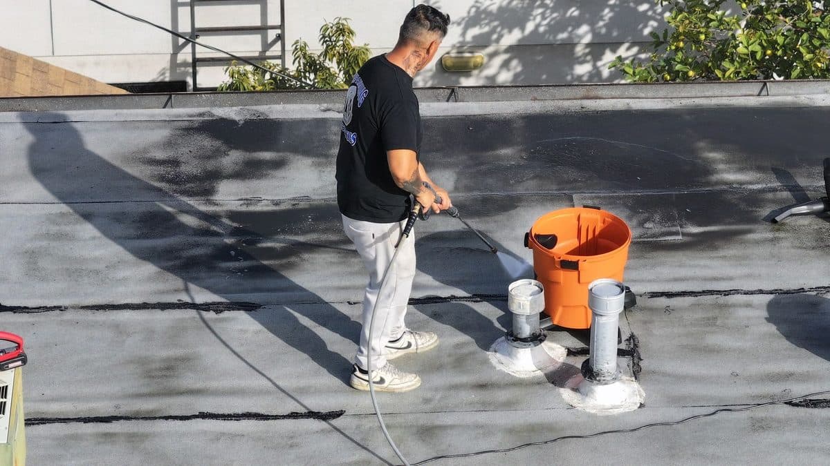 Crew member pressure washing a flat roof beside a bucket and two pipe vents