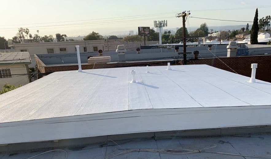 White flat roof with pipe vents and a raised edge, with neighboring buildings behind